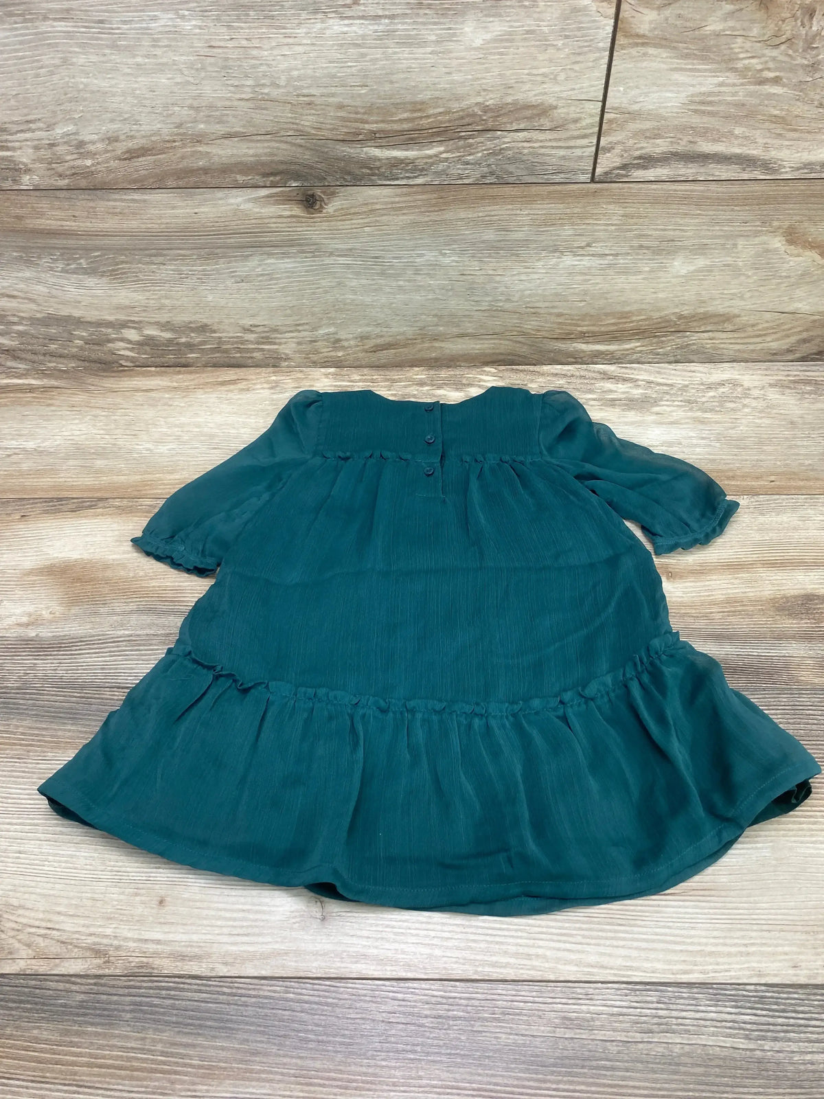 Top-down flat lay of green toddler dress showcasing back buttons, long puff sleeves, gathered waist, and ruffled hem on wooden floor