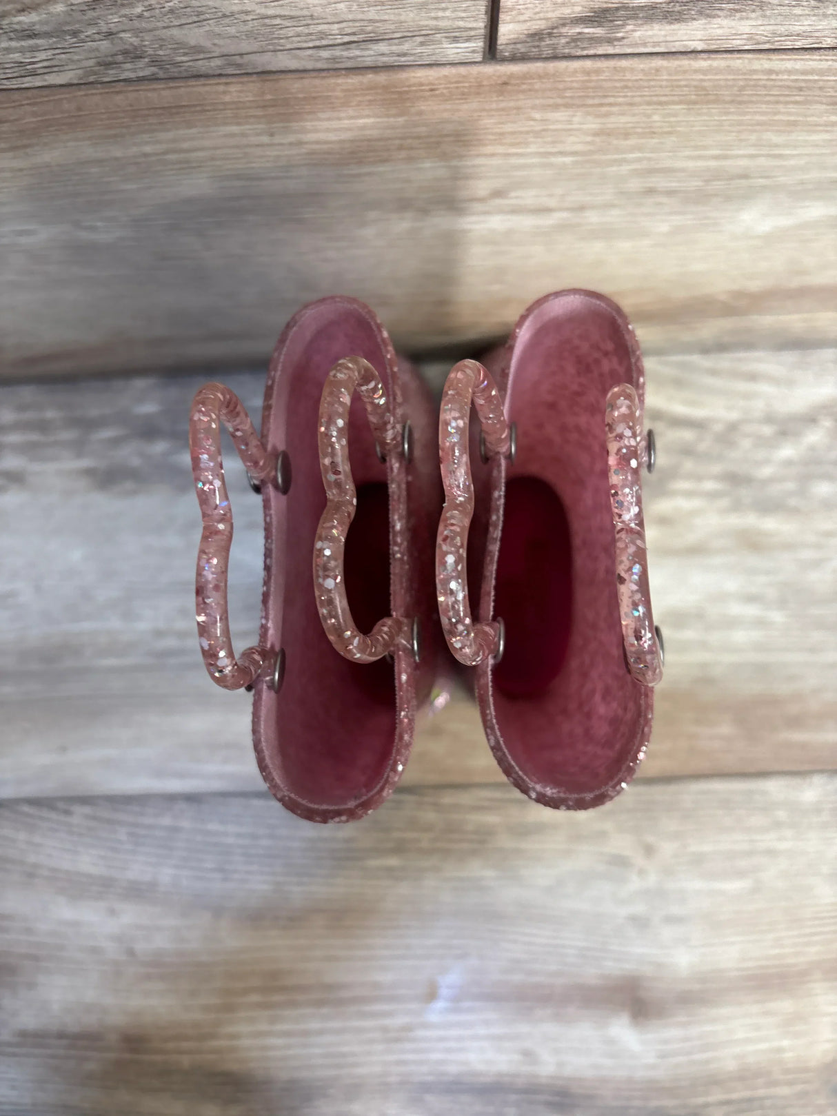 Top view of Western Chief Abby pink toddler rain boots showcasing glitter finish and wavy handles on wood floor