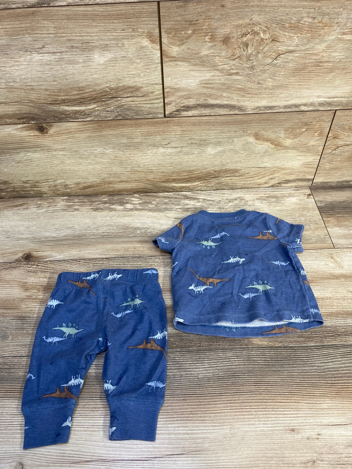 Flat lay of blue Child of Mine two-piece newborn shirt and pants set showing all-over dinosaur print on wood floor