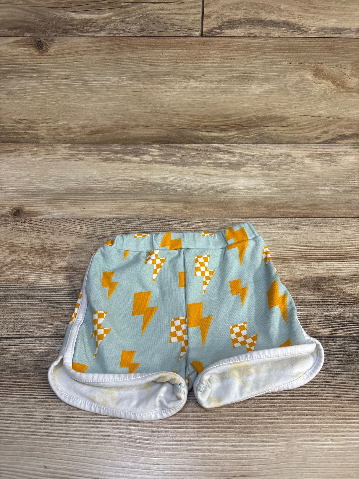 Flat lay of toddler lightning-print shorts showcasing yellow lightning bolts on light blue fabric with wood floor background