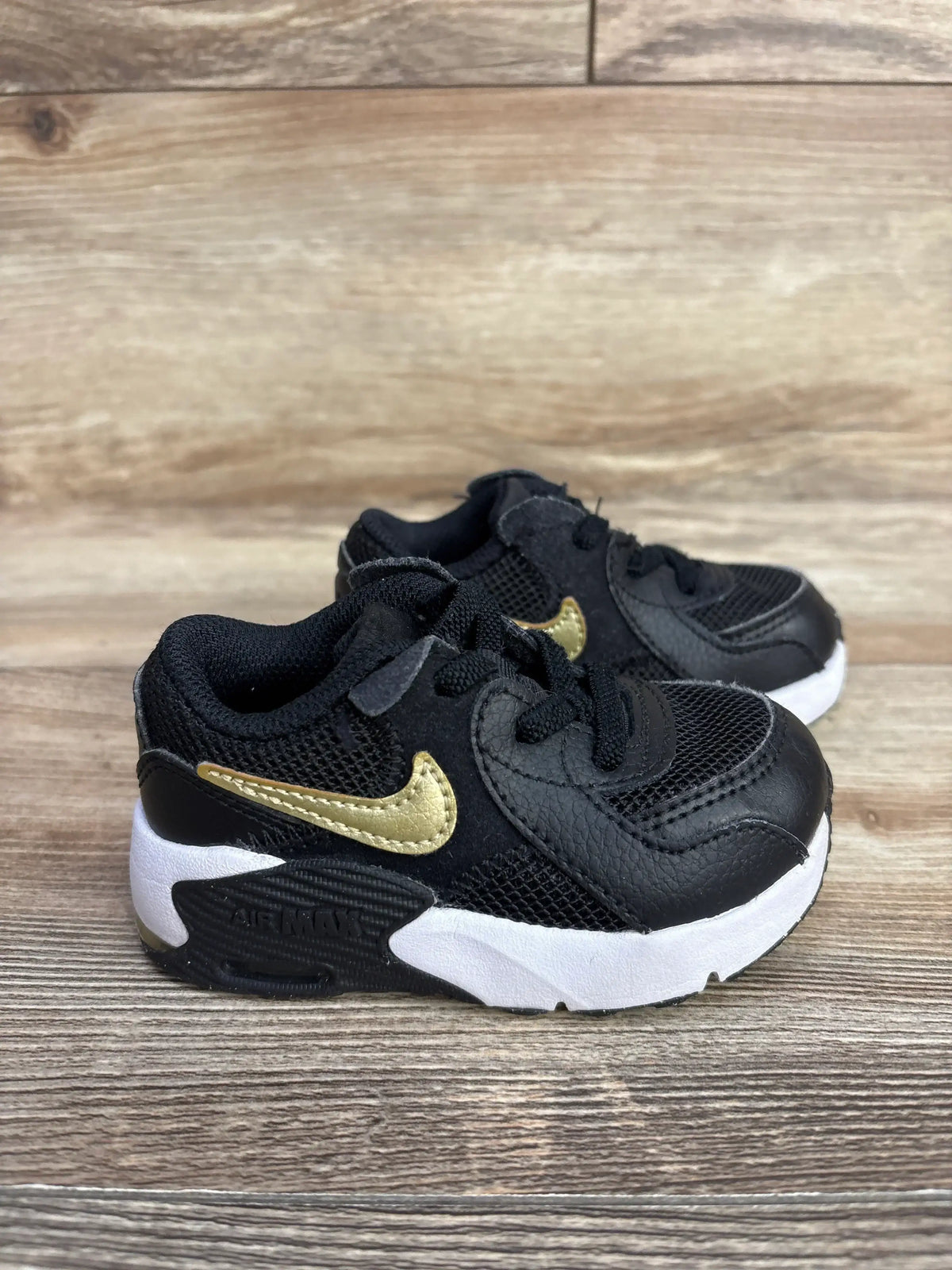 Black and gold Nike Air Max toddler sneakers with white soles on wooden surface