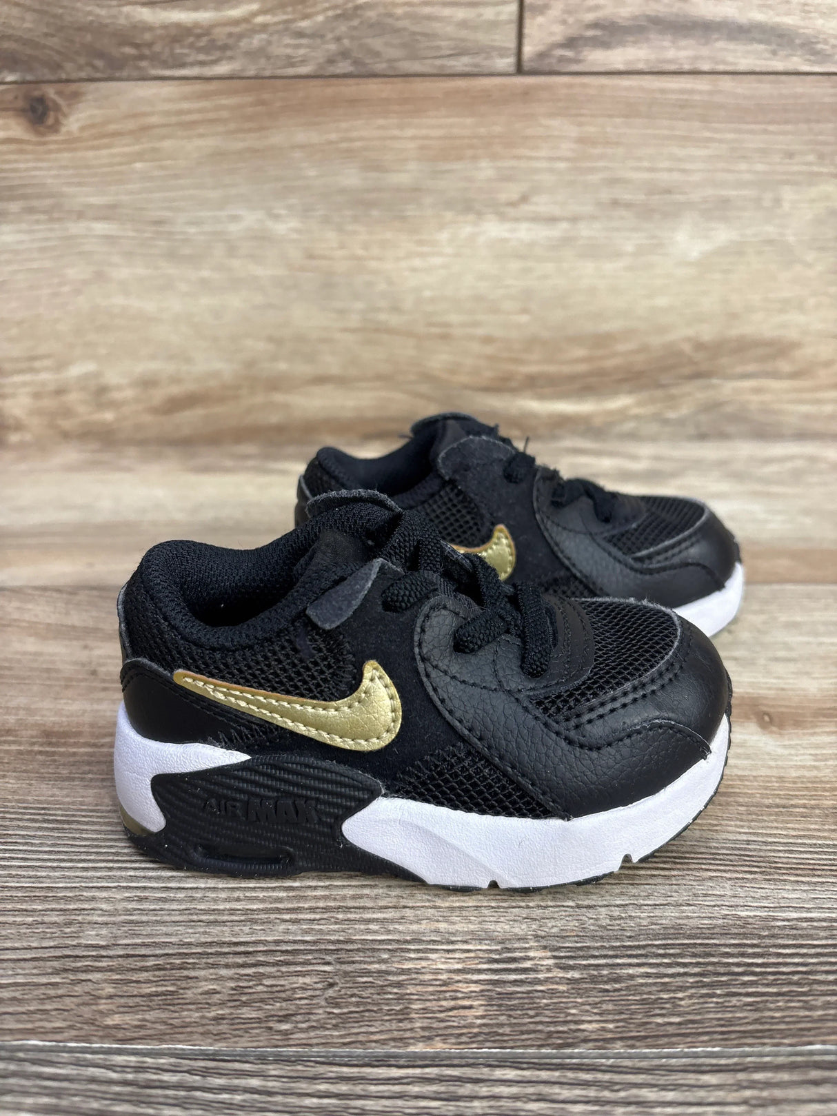 Black and gold Nike Air Max toddler sneakers with white soles on wooden surface