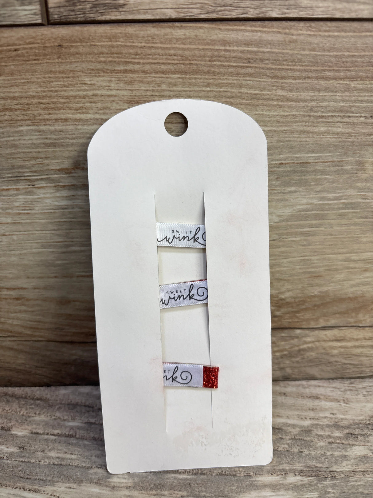 Front view of 3 Sweet Wink hair clips: two white ribbon clips with label and one with red glitter tip on white card over wood