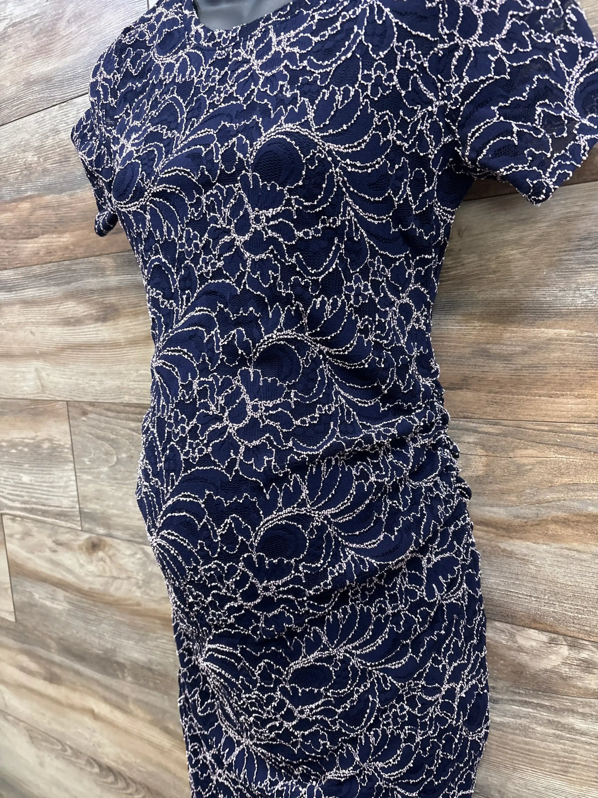Three-quarter view of navy Motherhood Maternity lace dress with embroidered overlay and side ruching on wood background