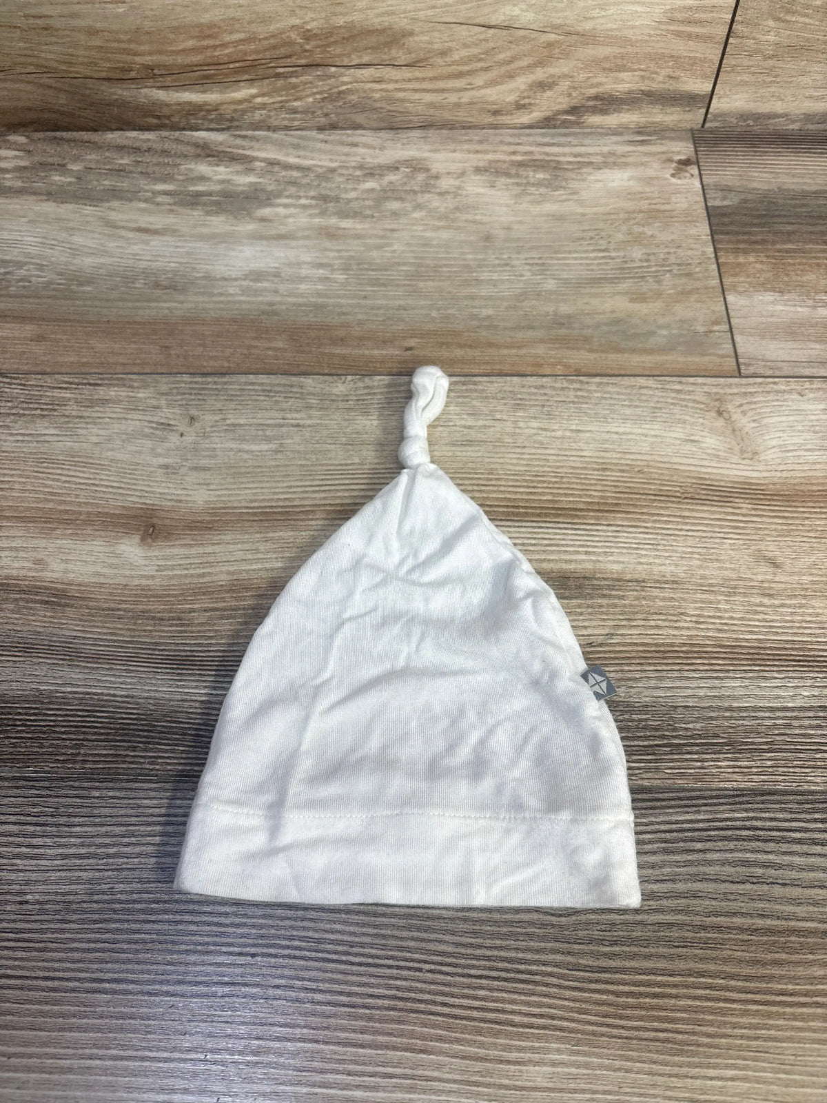 Front view of Kyte Baby white knotted cap in newborn size showcasing adjustable top knot and folded brim on a wood grain surface