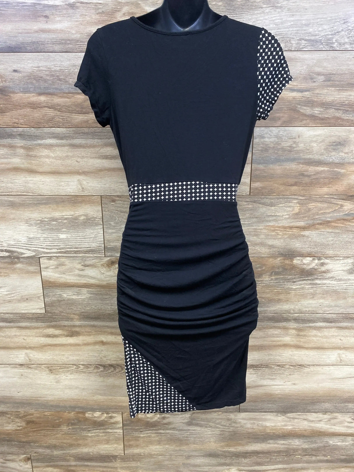 Front view of Kimi + Kai black maternity bodycon dress showing ruched skirt and patterned trim with wood background