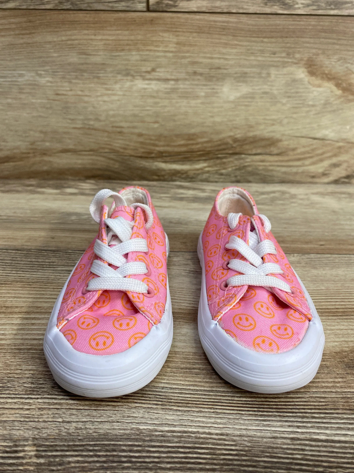 Pair of pink children's sneakers with orange smiley face patterns and white laces on wooden surface