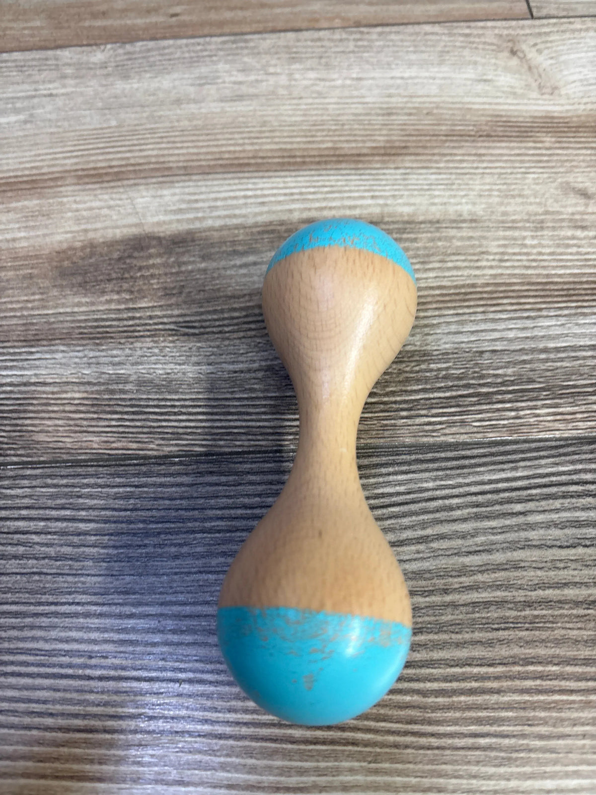 Top view of Lovevery wooden rattle showcasing turquoise ends and wooden finish on a wood grain surface