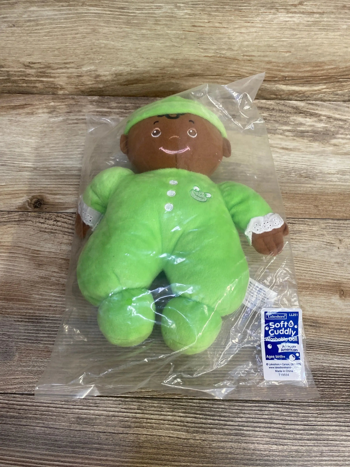 Front view of Lakeshore cuddly washable baby doll in bright green sleeper, brown plush skin, embroidered smile and lace cuffs inside clear plastic bag on wood surface