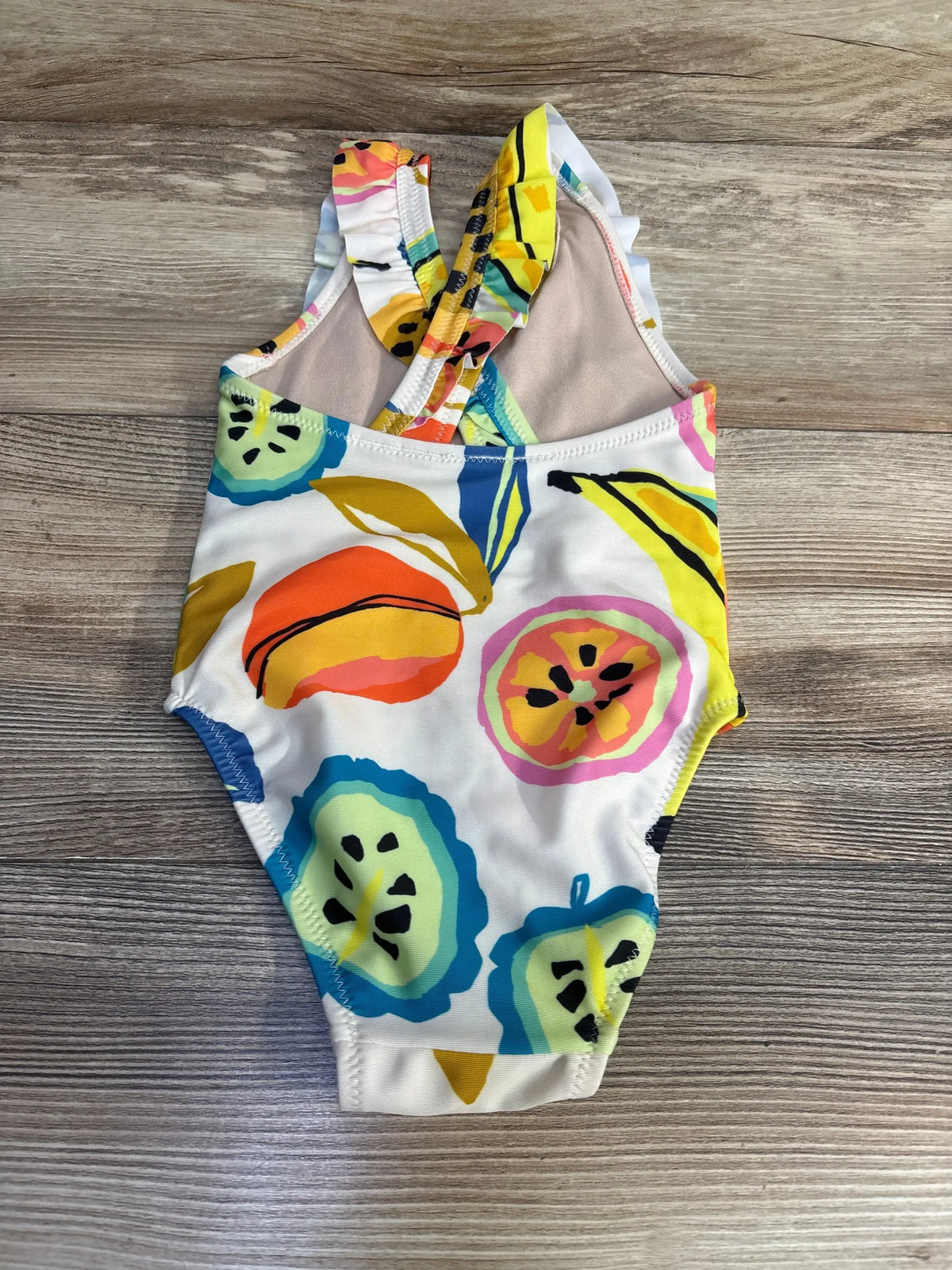 Flat lay of white toddler one-piece fruit-print ruffle swimsuit showing crisscross ruffle straps on wood background