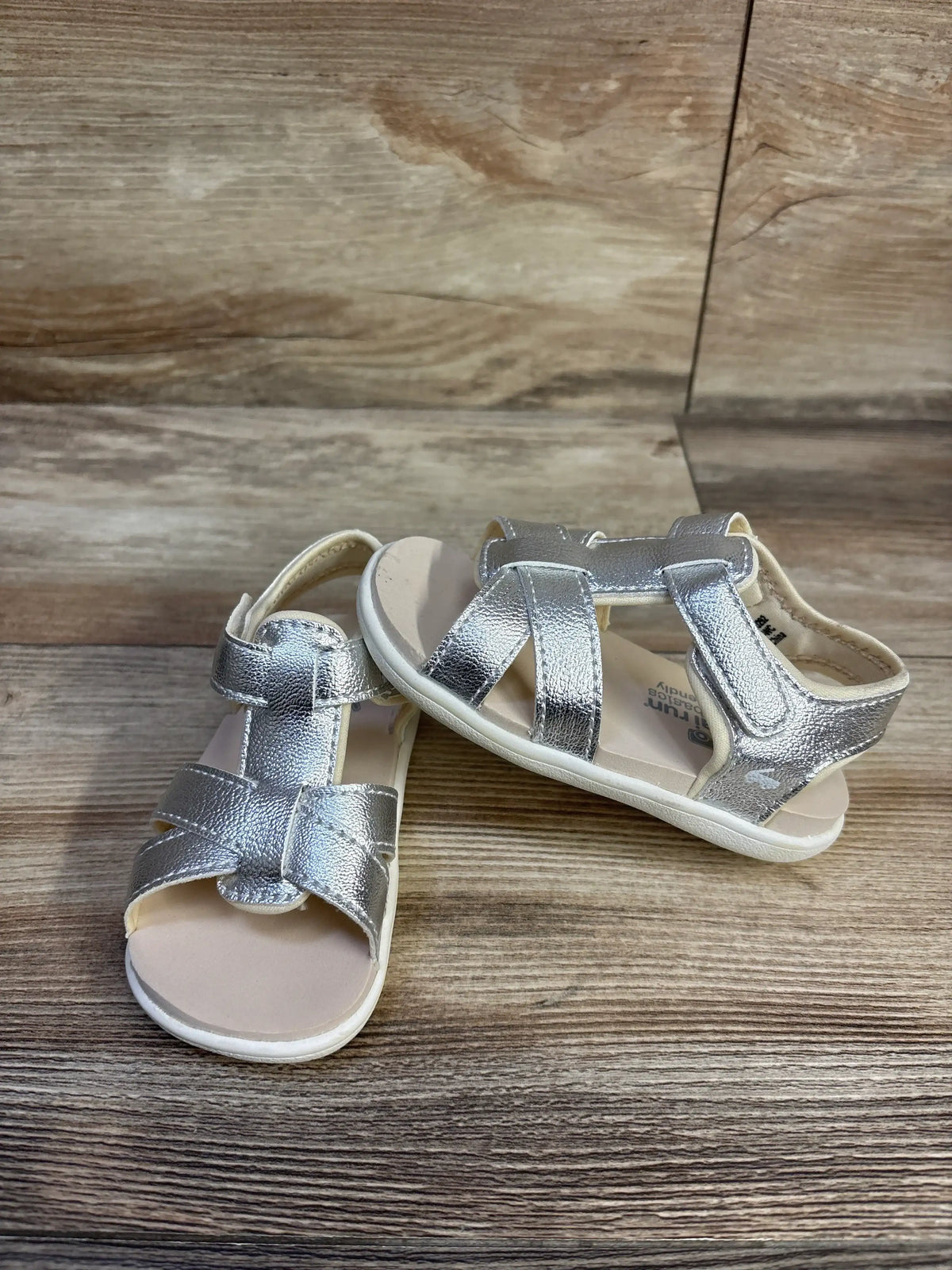 Front and side views of See Kai Run Shayna sandals in silver metallic with adjustable straps on a wood surface