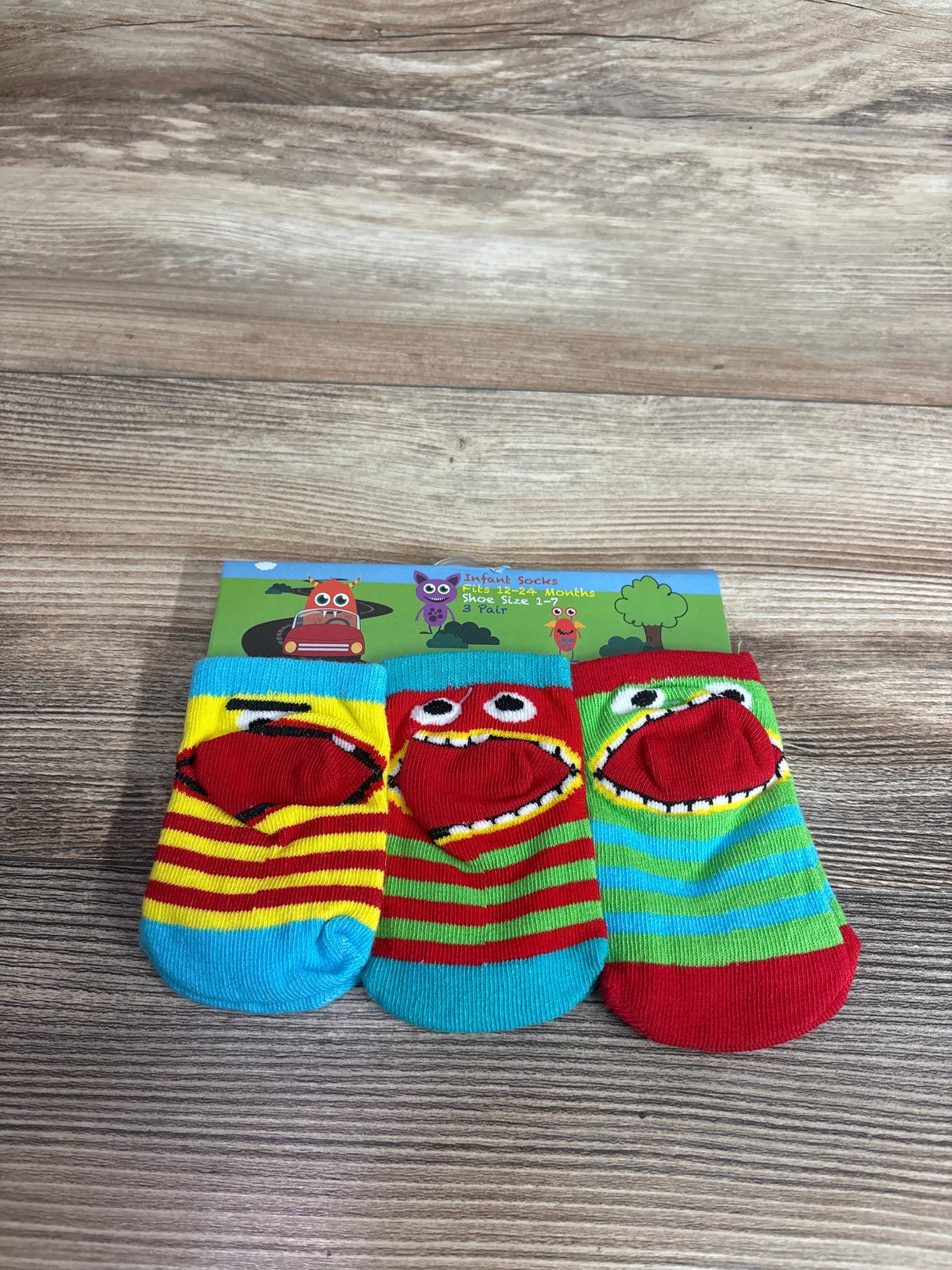 Front view of 3-pack baby socks showcasing colorful striped monster-face designs with cartoon header on wood background