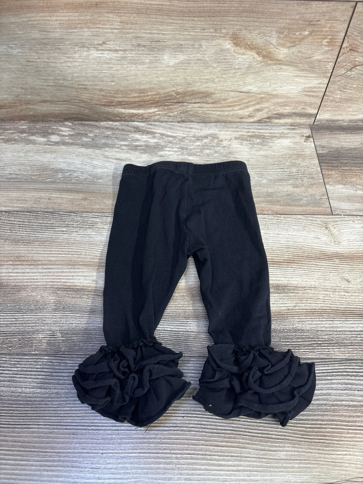 Top view of black Ruffled Rose leggings in size 12 months showcasing layered ruffled cuffs on a wood-look floor