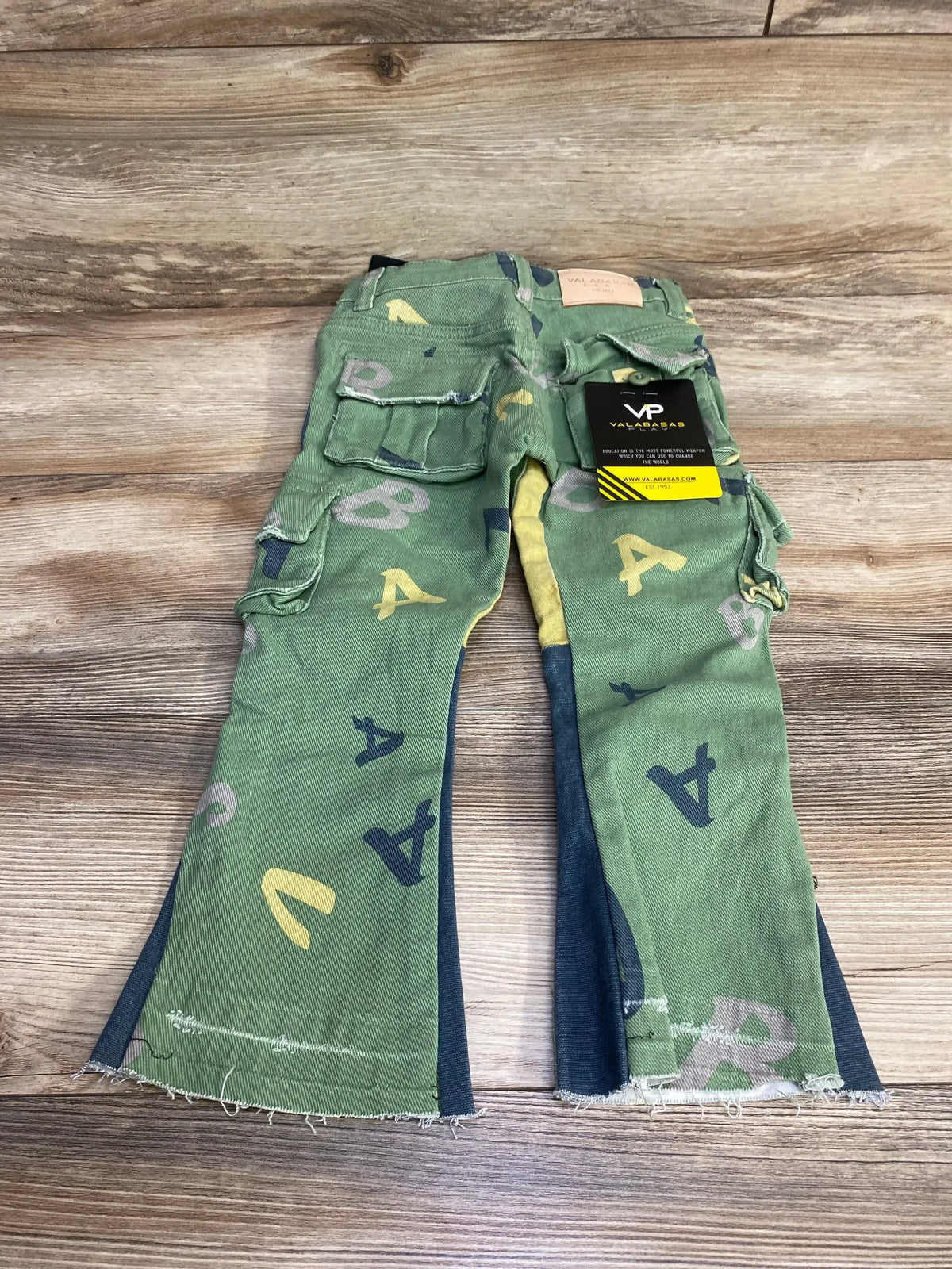 Flat lay of green Valabasas cargo pants with letter print, cargo pockets, dark side panels and frayed hems on wood
