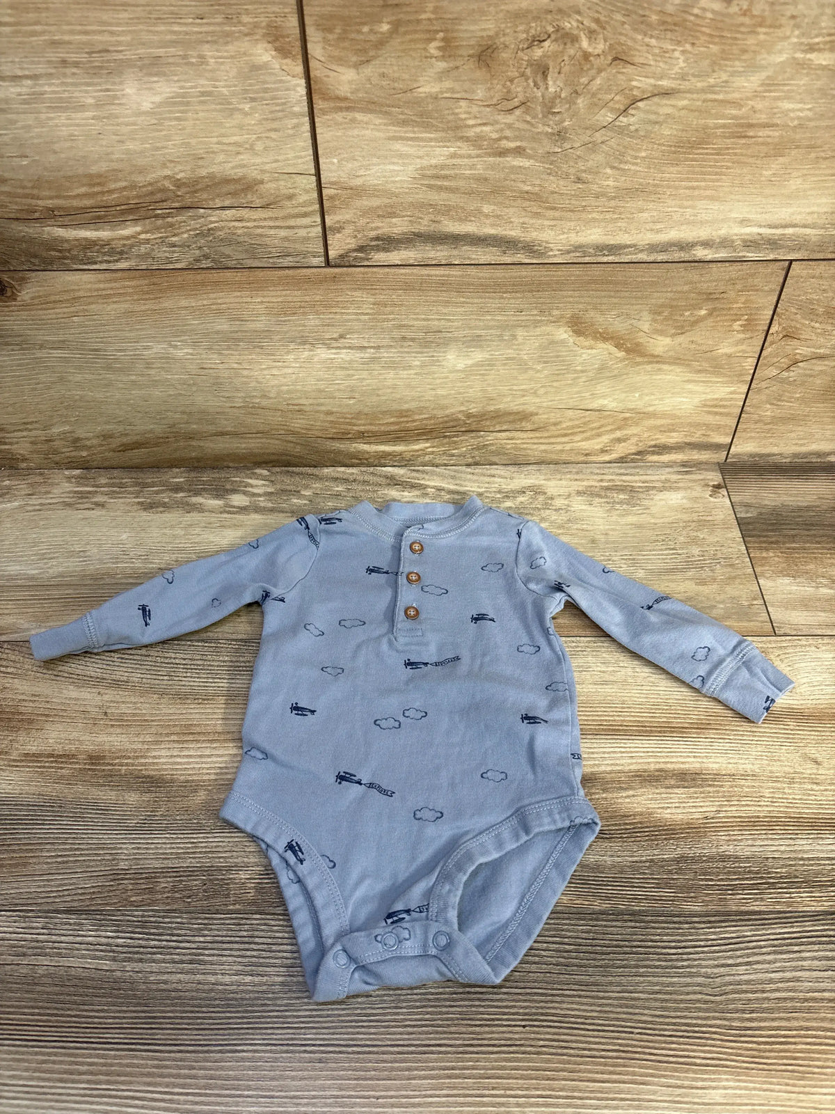Front view of Just One You blue henley bodysuit in size 6 months with cloud and airplane prints on a wood-look floor