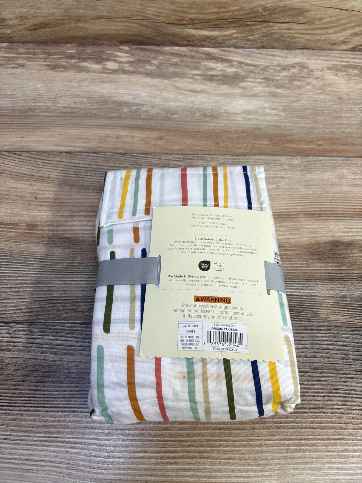 Folded Cloud Island fitted crib sheet showing multicolored dash stripes and product label on wood surface