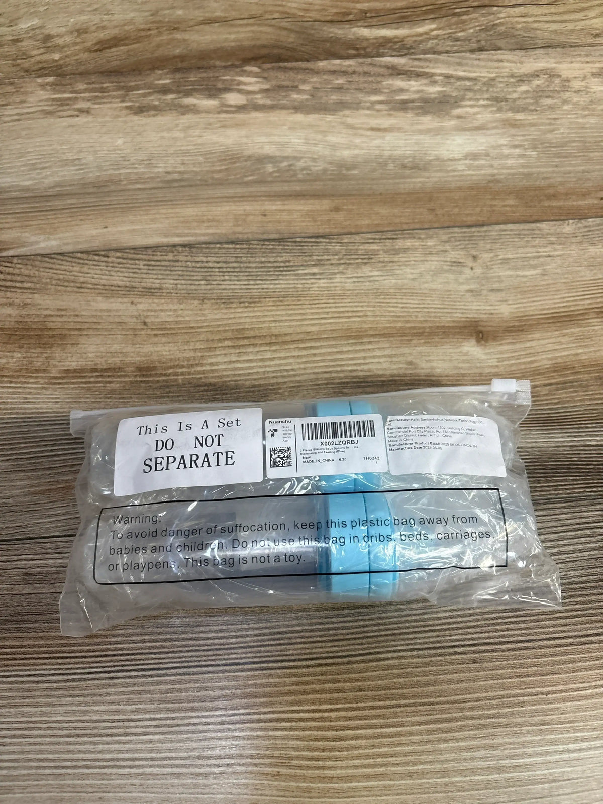 Top-down view of Nuanchu blue silicone baby spoons 2-pack in sealed clear plastic bag showing large label This Is A Set DO NOT SEPARATE on a wood surface