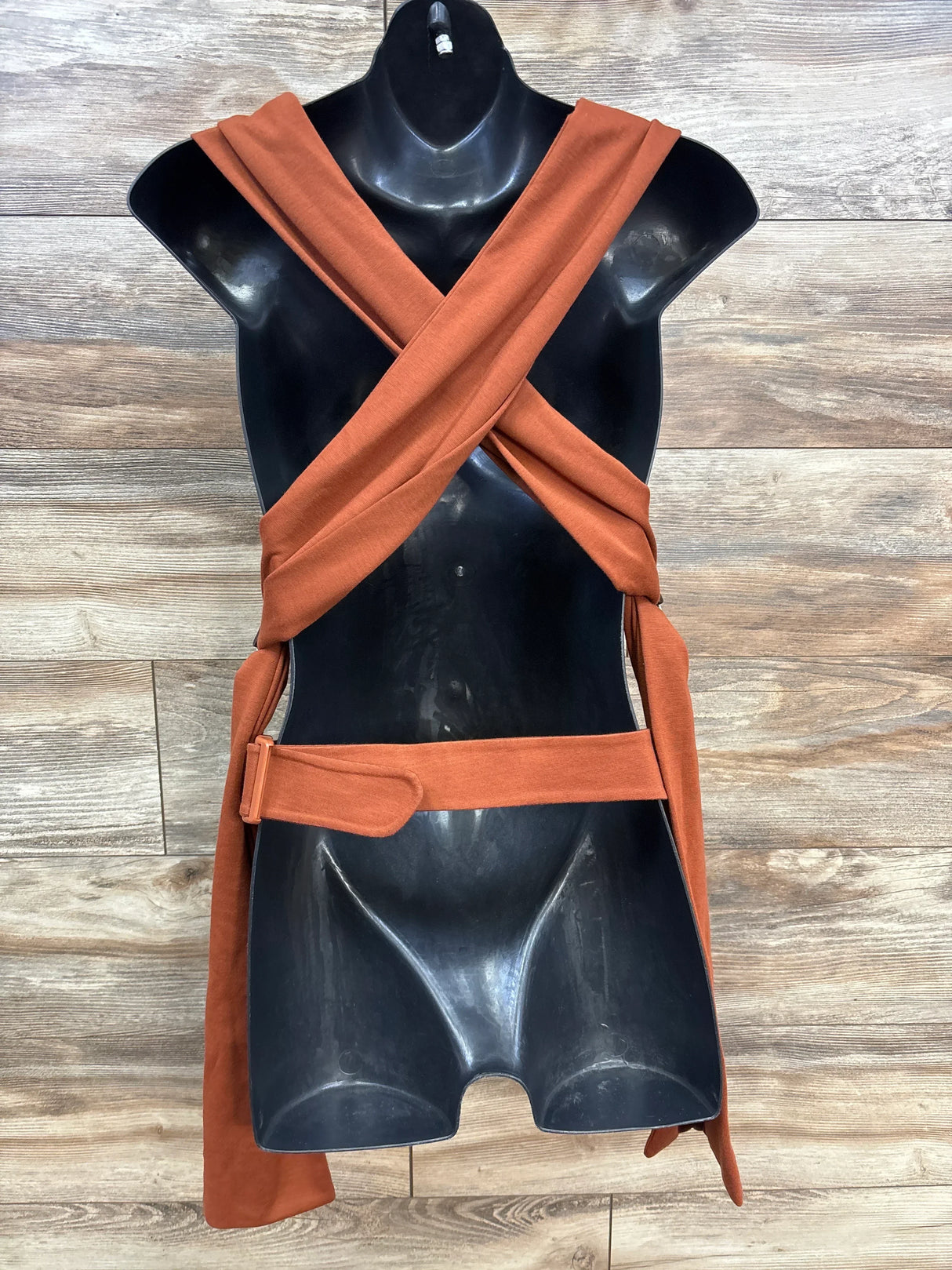 Black mannequin wearing rust orange wrap dress with cross front straps and belt on wooden floor
