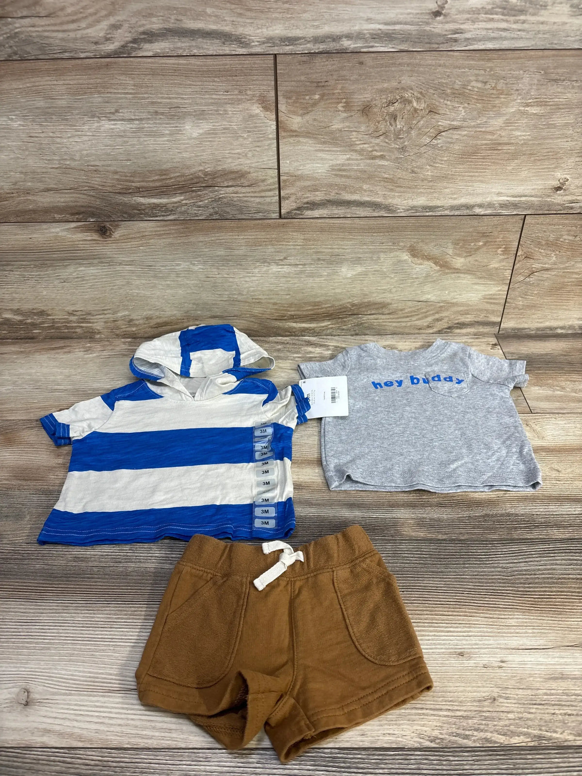 Flat lay of 3-piece baby set size 3M showing blue-and-white hooded shirt, grey hey buddy tee and brown shorts on wood background