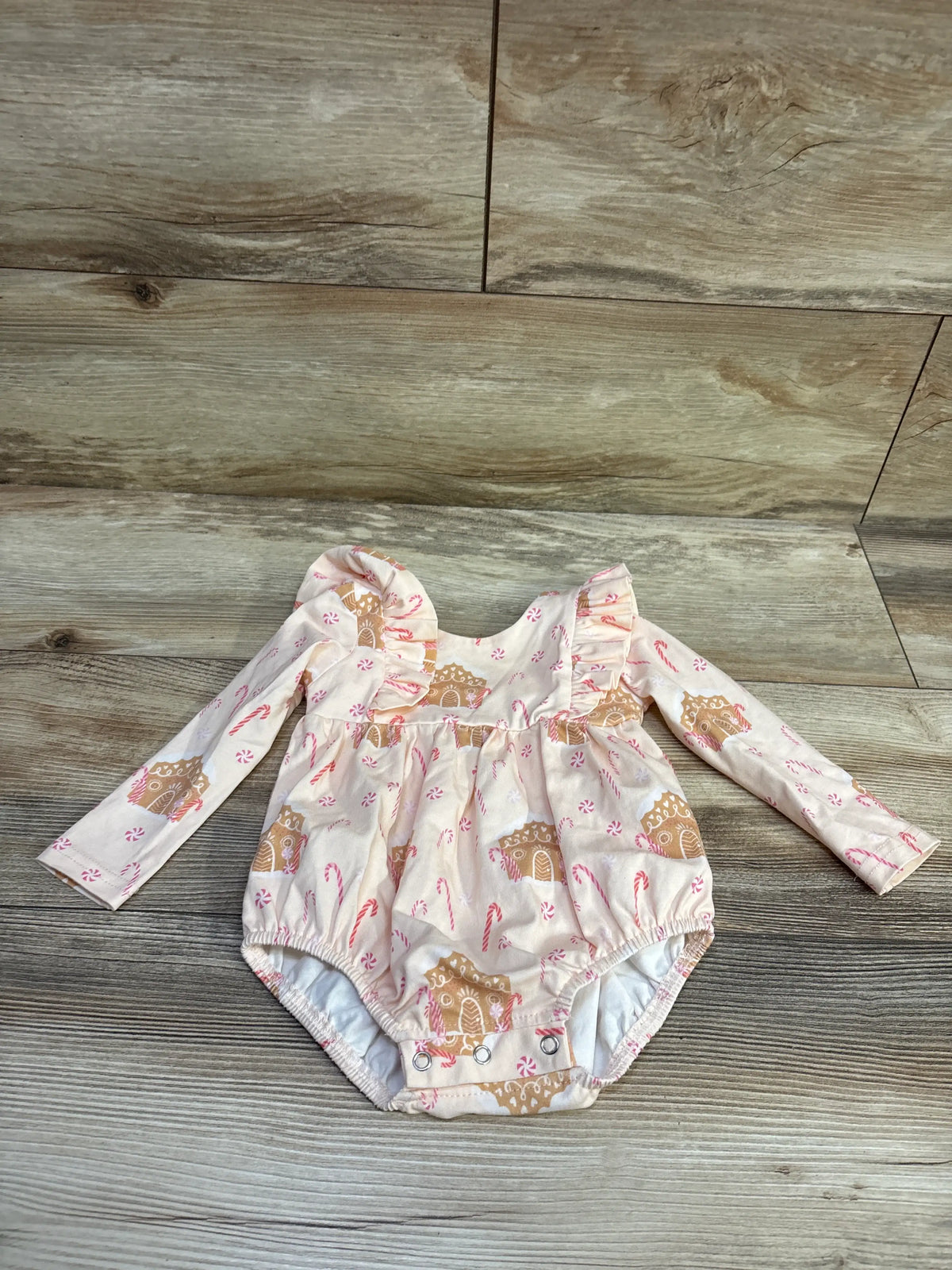 Front view of cream long-sleeve gingerbread house bubble romper showcasing ruffled shoulders and candy cane print on wood-look flooring