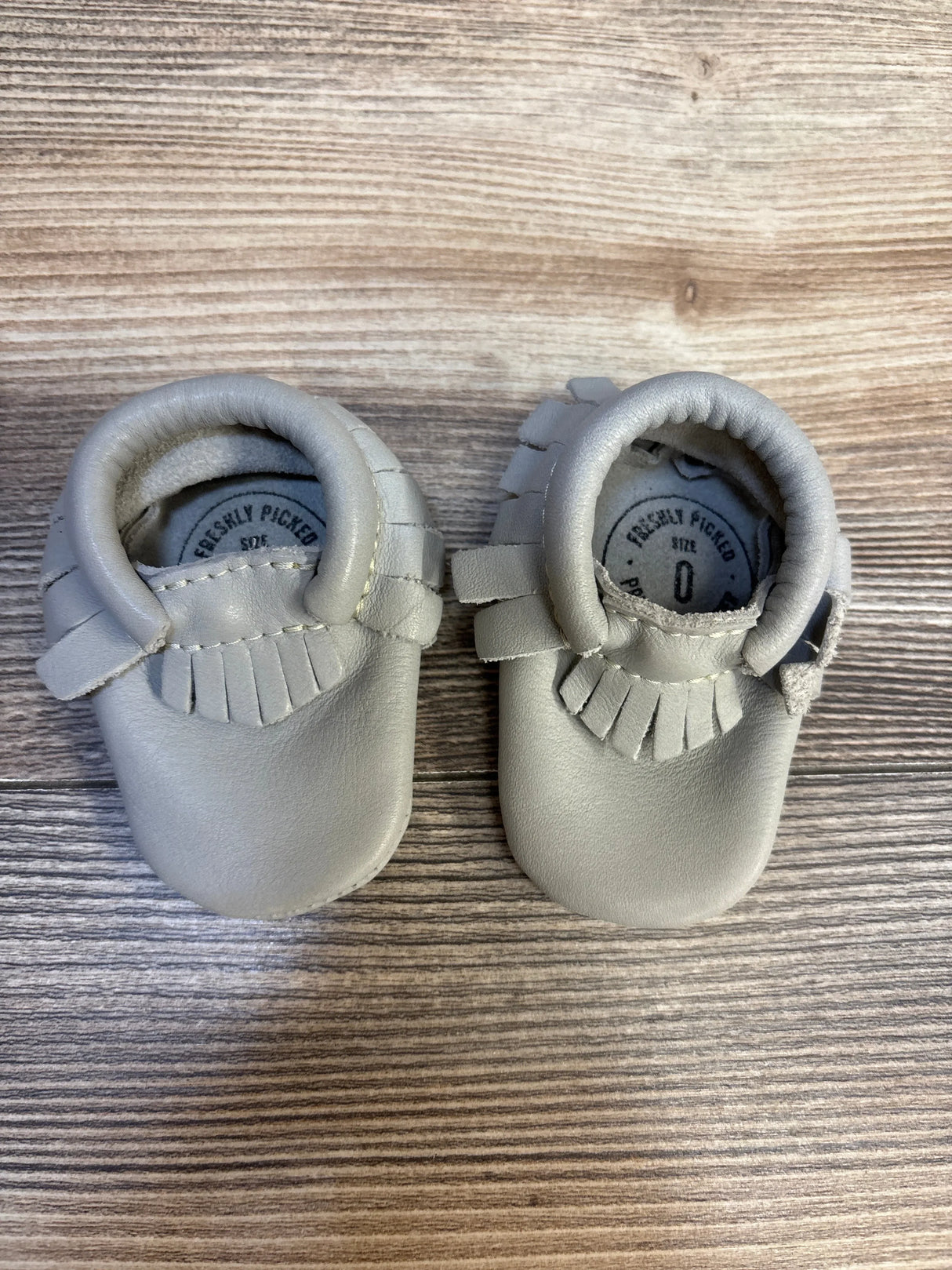 Gray leather baby moccasins with fringe detail on wood background, size 0 infant shoes