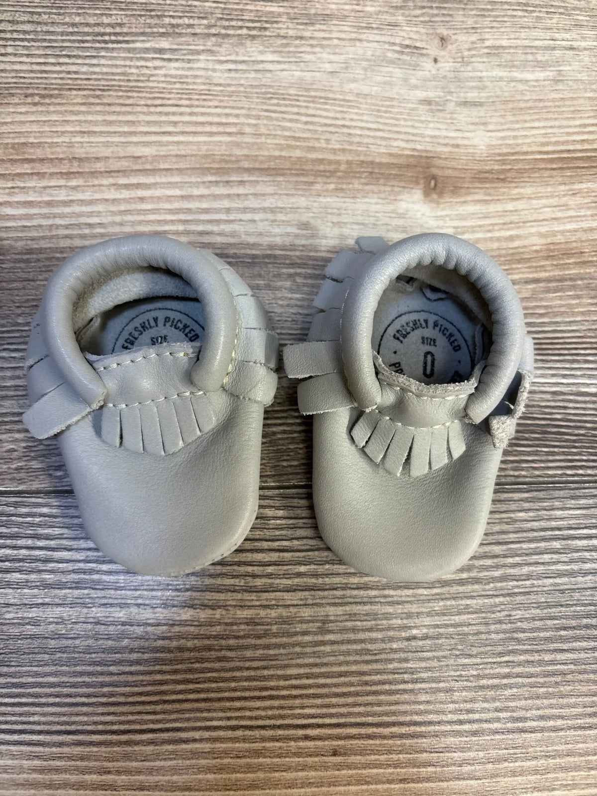 Gray leather baby moccasins with fringe detail on wood background, size 0 infant shoes