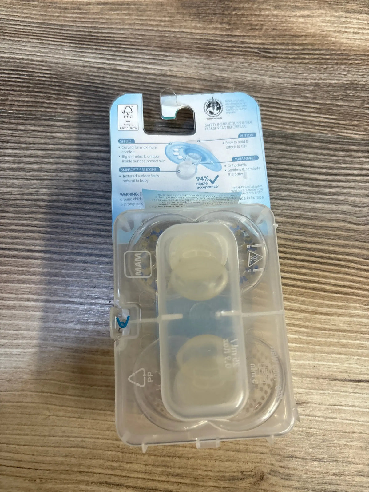 Top view of NEW MAM 2-pack clear pacifiers (16+ months) in translucent travel case showing two silicone nipples on wood