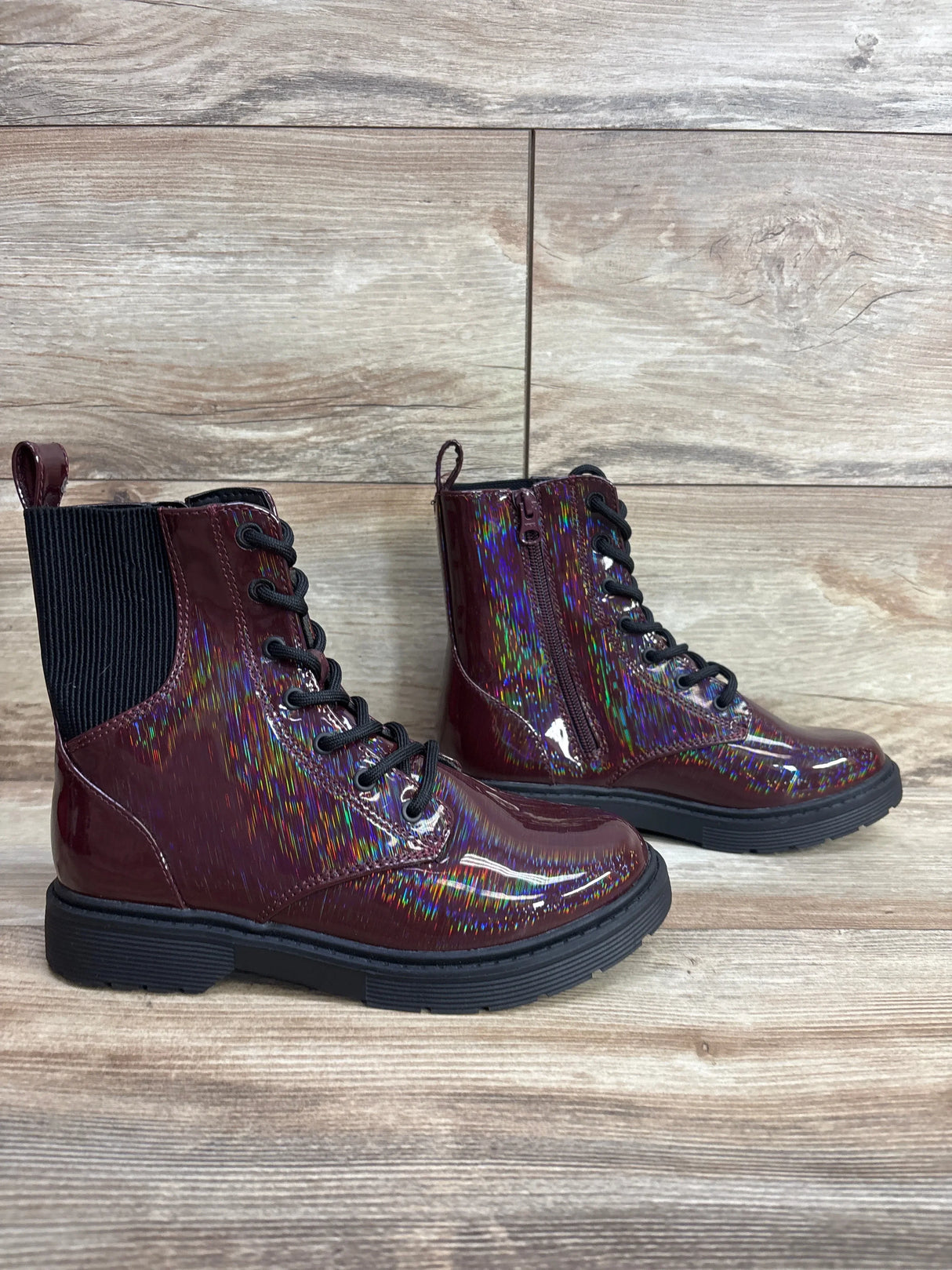 Burgundy patent leather kids lace-up boots with black soles and rainbow shine on wood floor