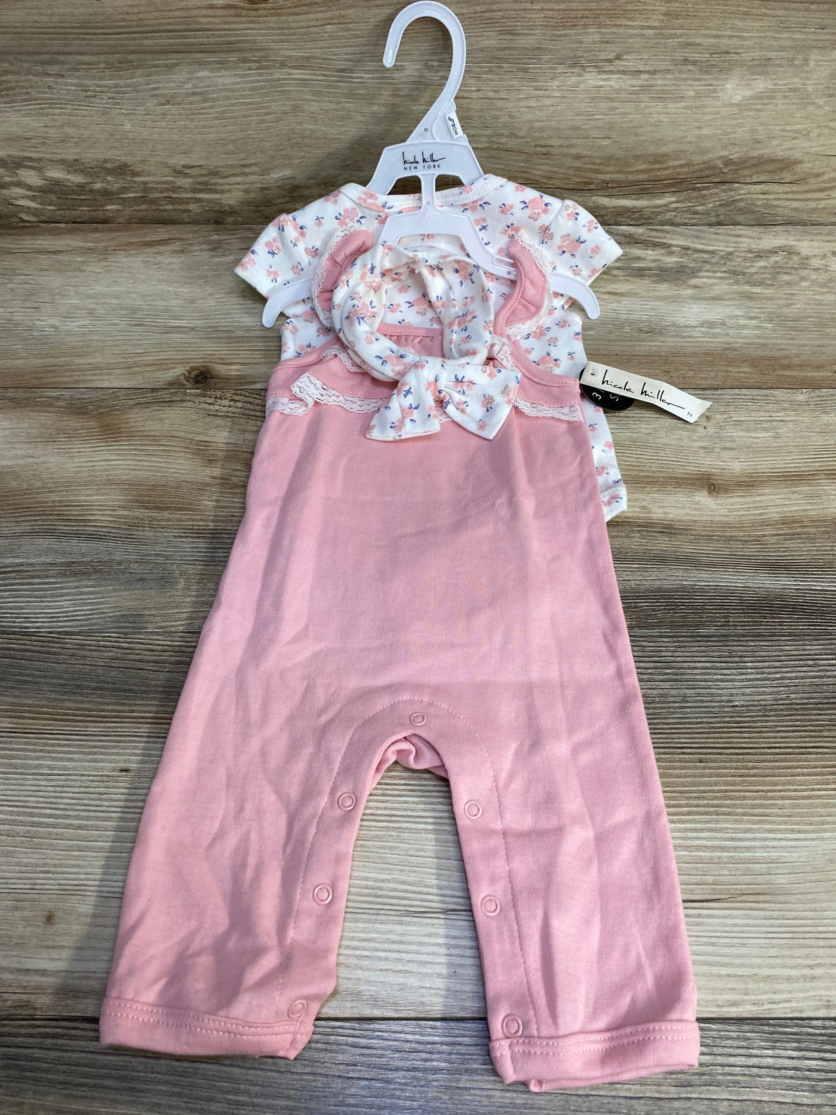 Front of Nicole Miller baby set: white floral bodysuit, pink jumpsuit with ruffle and leg snaps, matching headband on wood