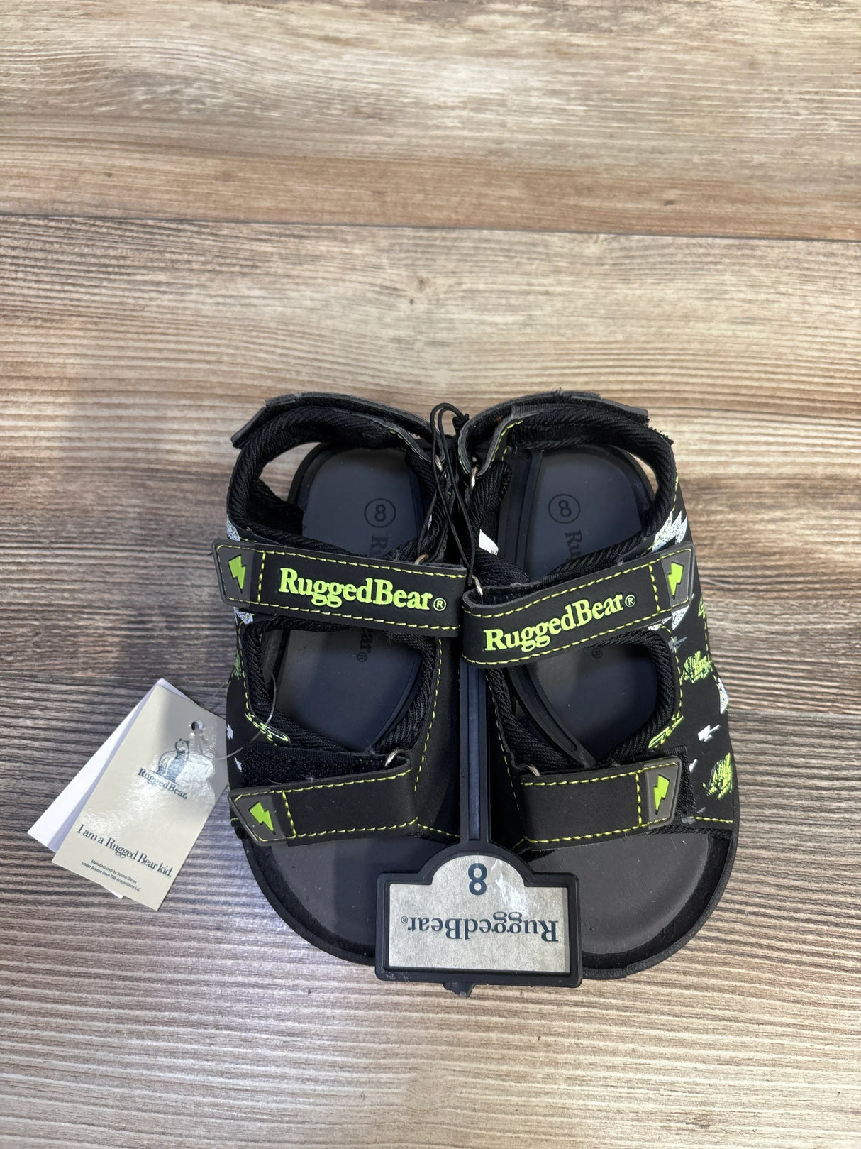 Top-down view of Rugged Bear black open-toe toddler sandals size 8C showing two Velcro straps with neon green RuggedBear logo and tag on wood floor