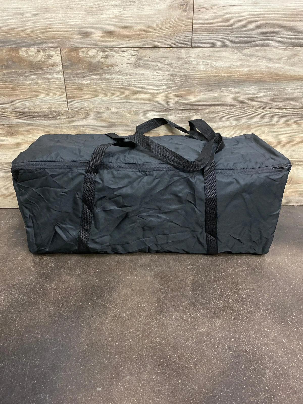 Front view of 4moms Breeze Go playard packed in dark grey zippered carrying bag showing shoulder straps against wood wall