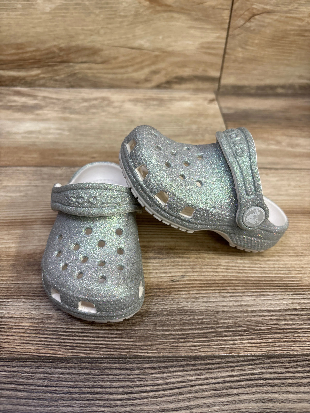 Front and side views of silver glitter Crocs Classic Clogs showcasing perforated uppers and heel straps on a wood-grain floor