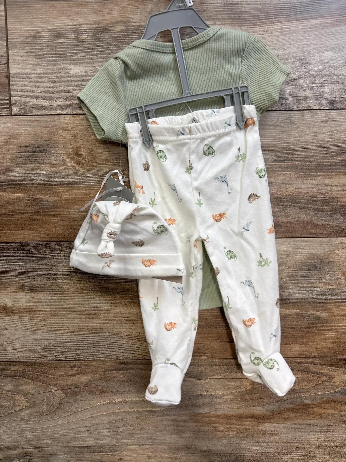 Front view of 3-piece baby set, ribbed sage-green bodysuit, white footed pants and knotted hat with dino print on wood