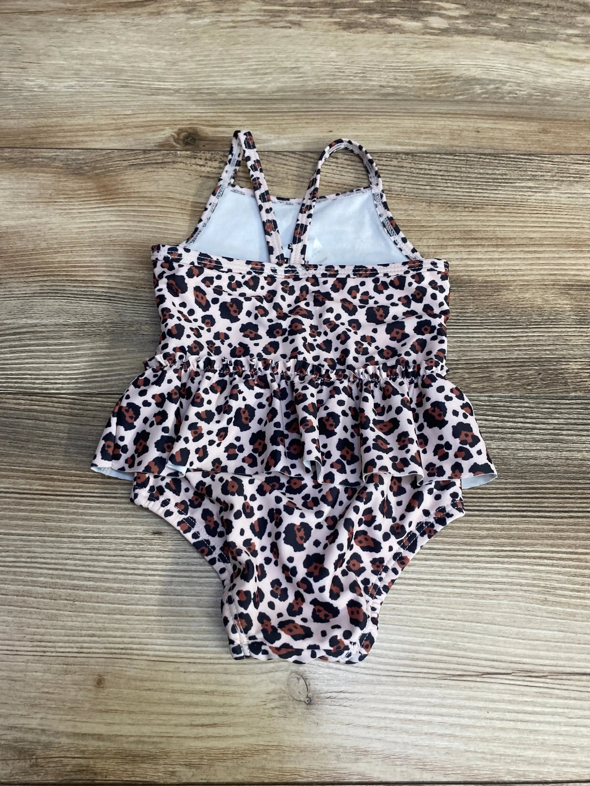 Top-down view of pink newborn one-piece leopard print swimsuit with crossed shoulder straps and ruffle peplum on wood background