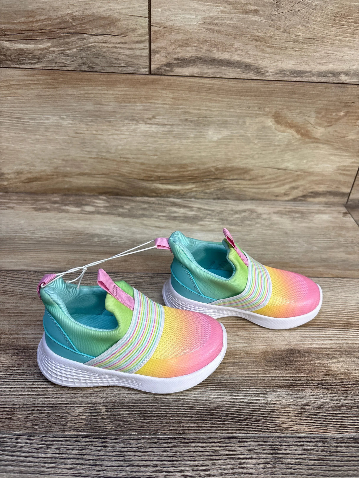 Angled top view of Cat and Jack toddler slip-on sneakers showing rainbow gradient upper, elastic striped band, and white cushioned sole on wood background