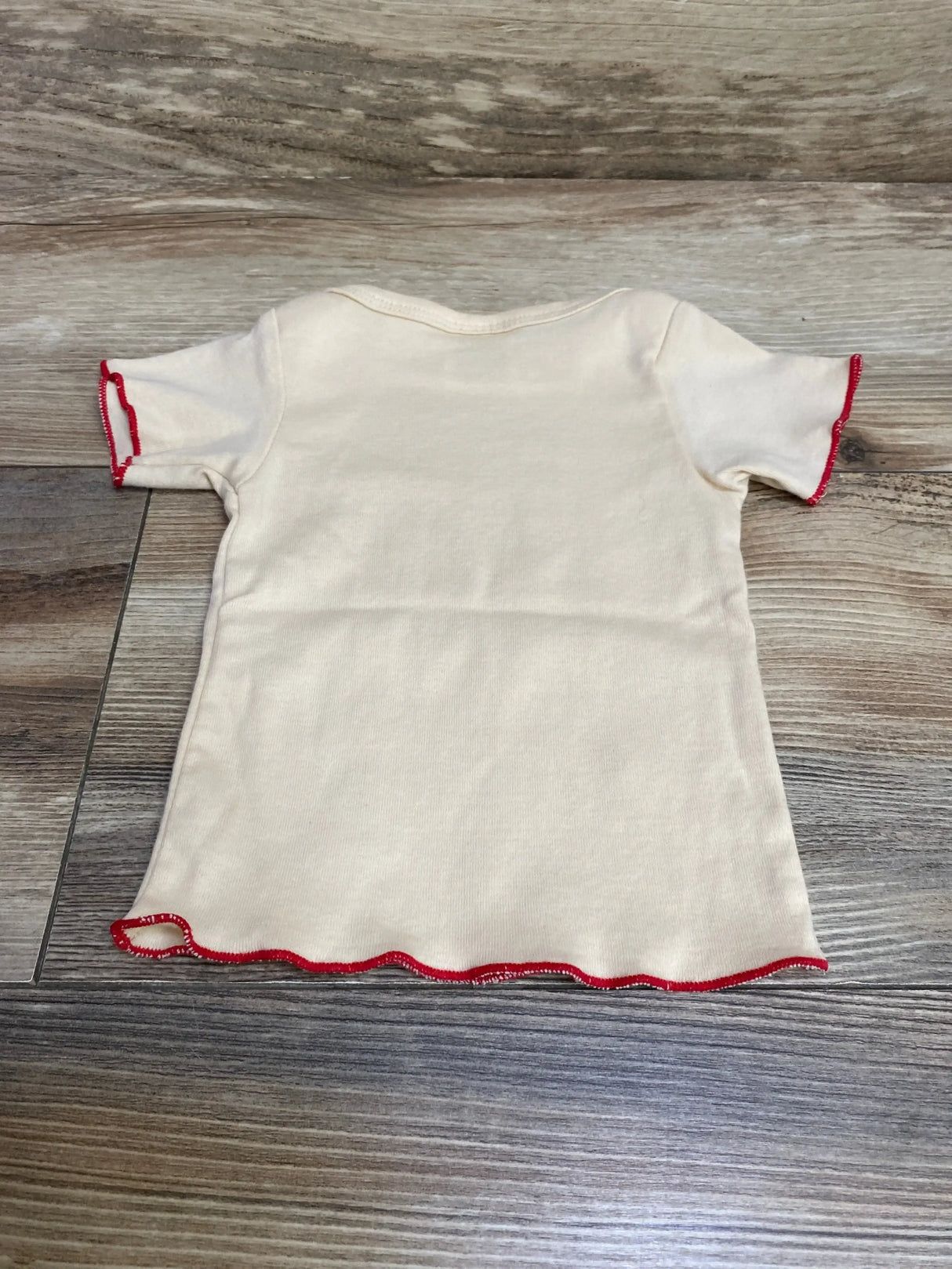 Back flat-lay of Wrybaby Mysterio cream 0-12m shirt showcasing red lettuce-edge hem and short sleeves on wood floor background