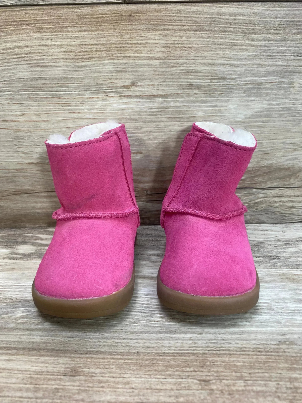 Front view of UGG Keelan toddler boots bright pink suede with faux fur lining and brown rubber soles on wood background