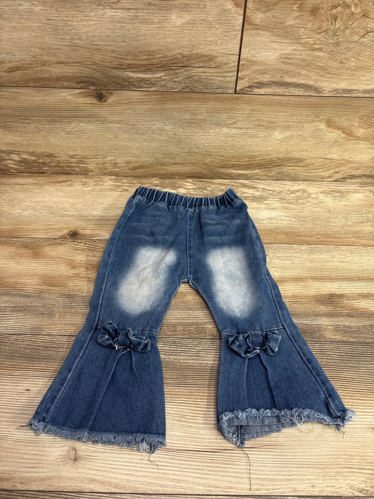 Front view of blue 12 to 18 months bell-bottom jeans with elastic waist, bows, faded knees, and frayed hems on wood flooring