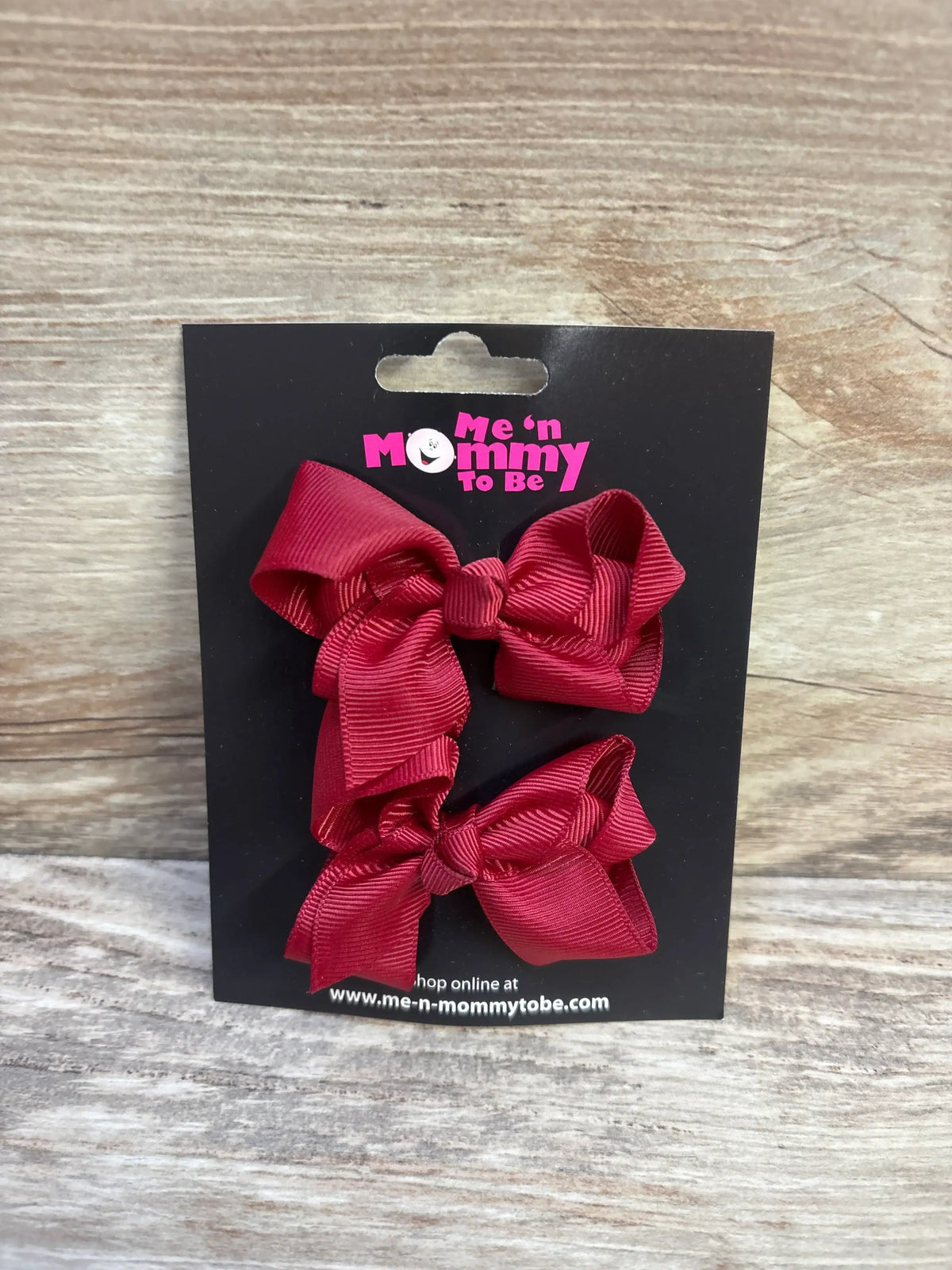 Front view of two maroon grosgrain baby hair bows showcasing ribbed ribbon texture on a black display card with wood tabletop background