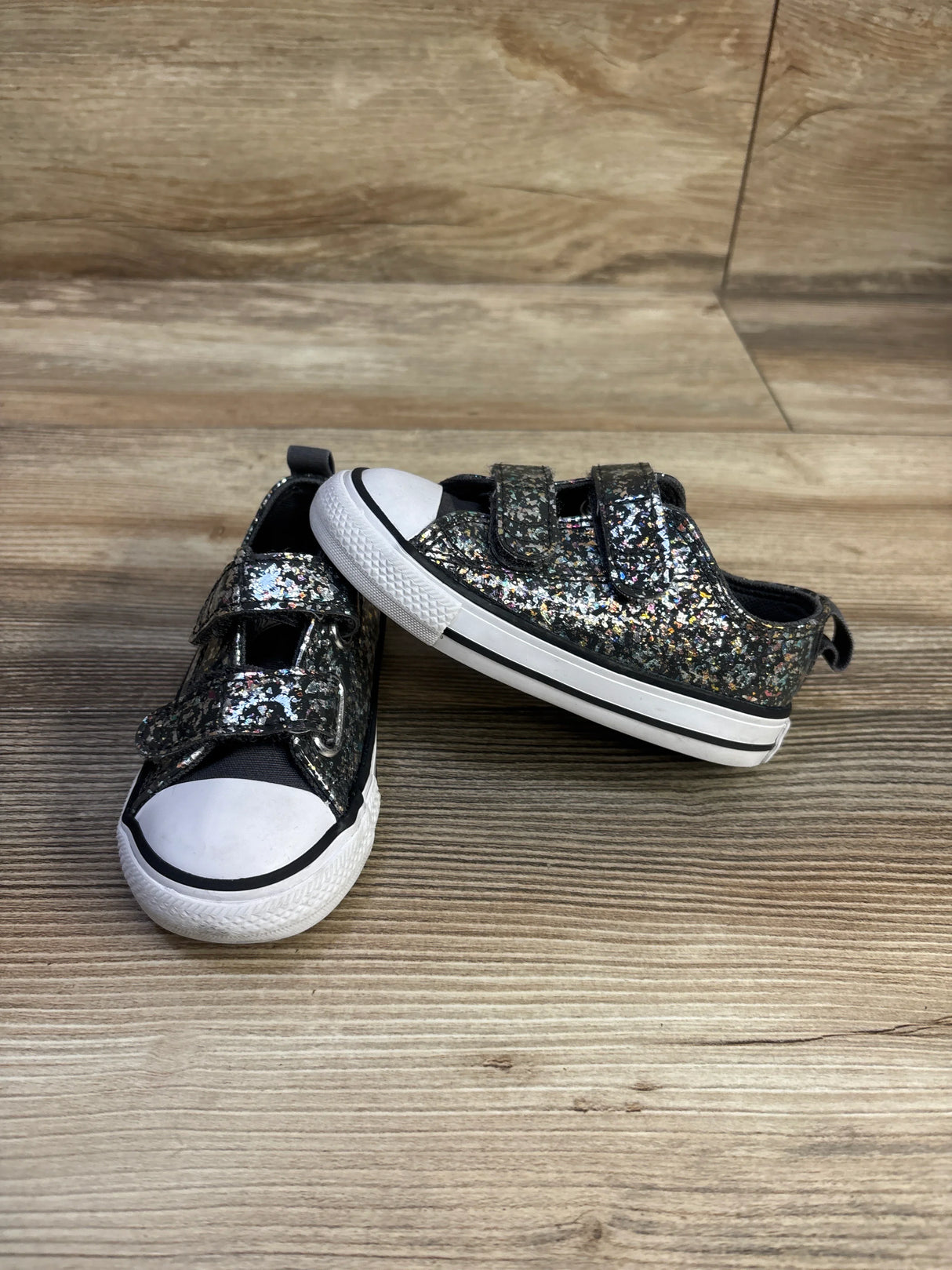 Angled view of Converse Chuck Taylor toddler sneakers with black glitter upper, white toe caps, and Velcro straps on wood