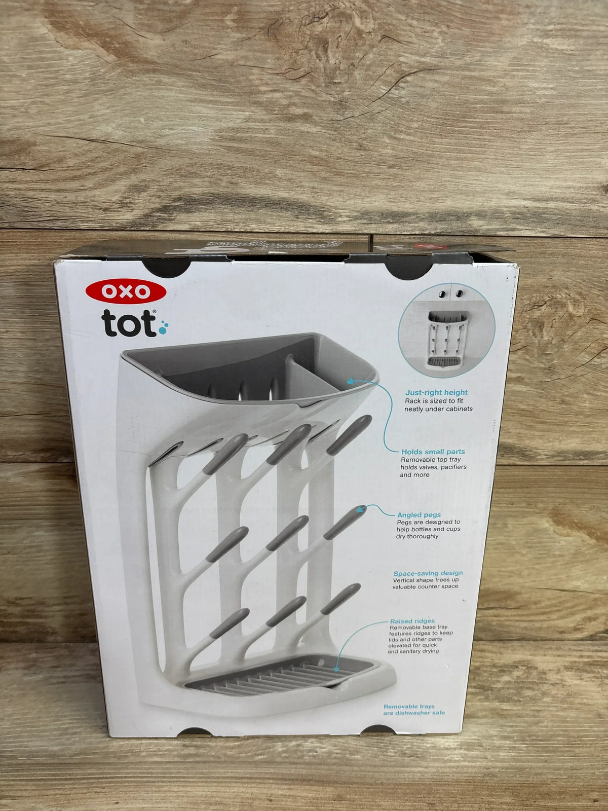 Front view of OXO Tot space-saving drying rack box showing angled vertical pegs and removable drip tray on wooden background