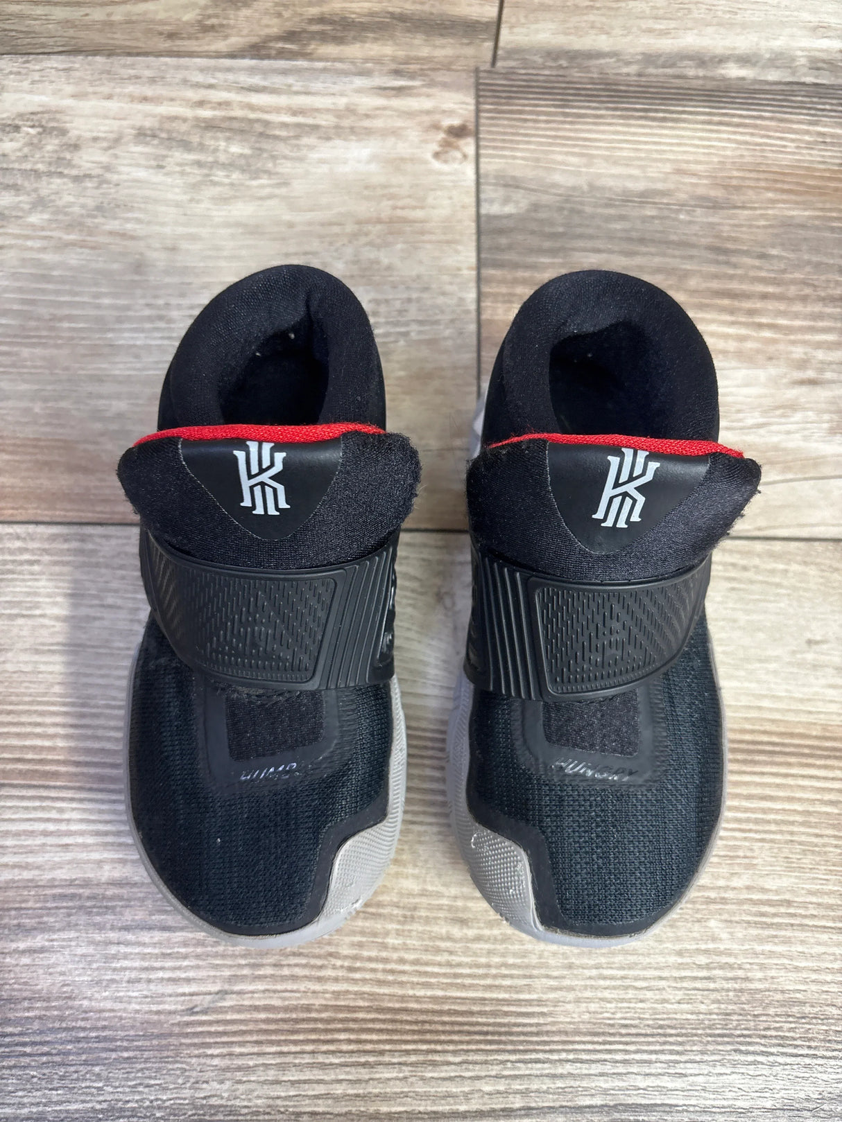 Top view of Nike Kyrie 6 TD Bred toddler sneakers showing black upper, red collar trim and Kyrie logo on wood floor