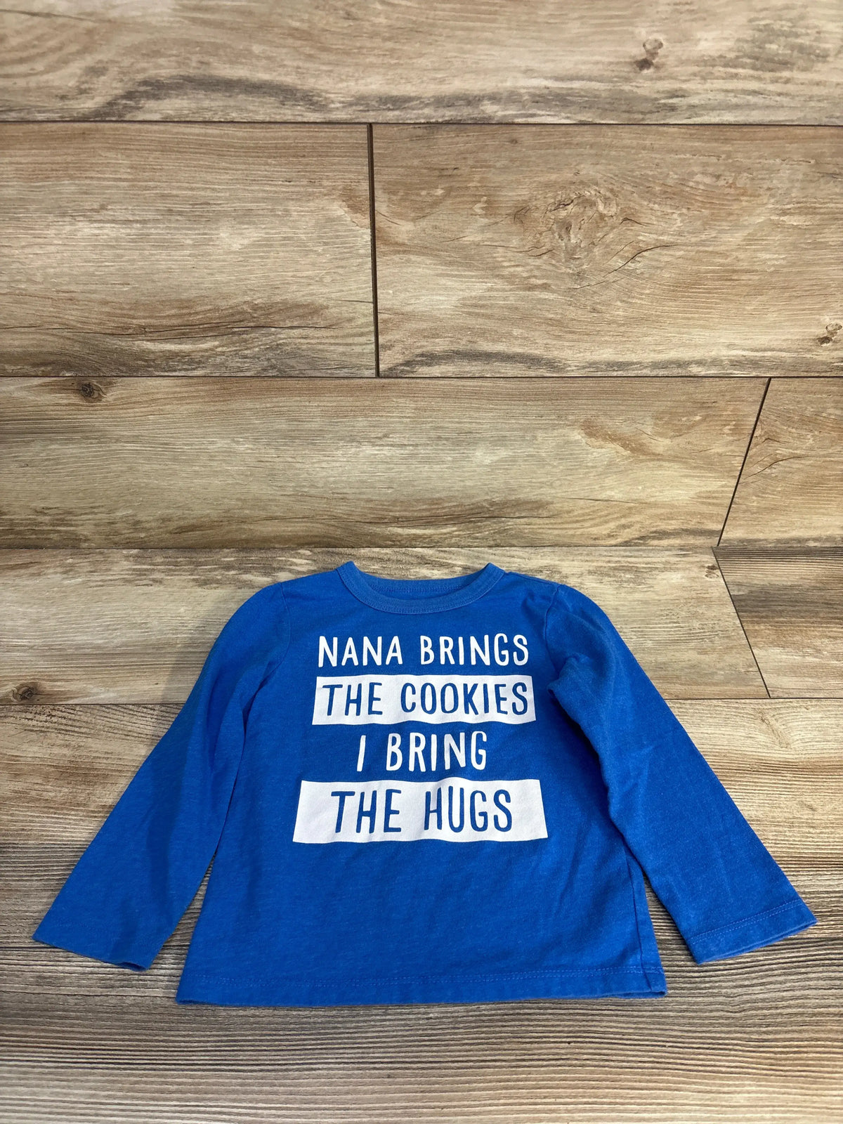 Front view of blue toddler long-sleeve shirt with Nana Brings The Cookies I Bring The Hugs graphic on wood floor