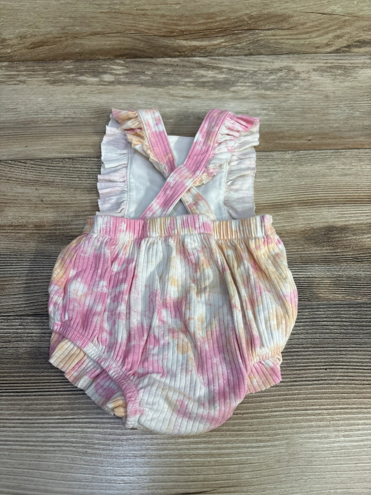 Flat lay of pink tie-dye baby romper (3-6 months) showing crossed ruffle shoulder straps, ribbed knit fabric, and elastic leg openings on a wood background
