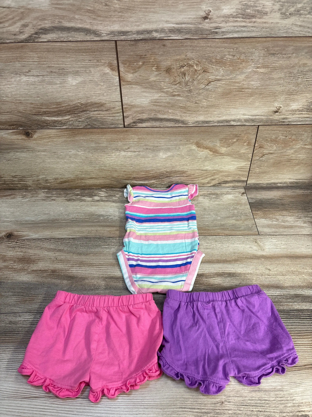 Top-down view of Children’s Place three-piece set showcasing striped bodysuit and pink and purple ruffled shorts on wood floor