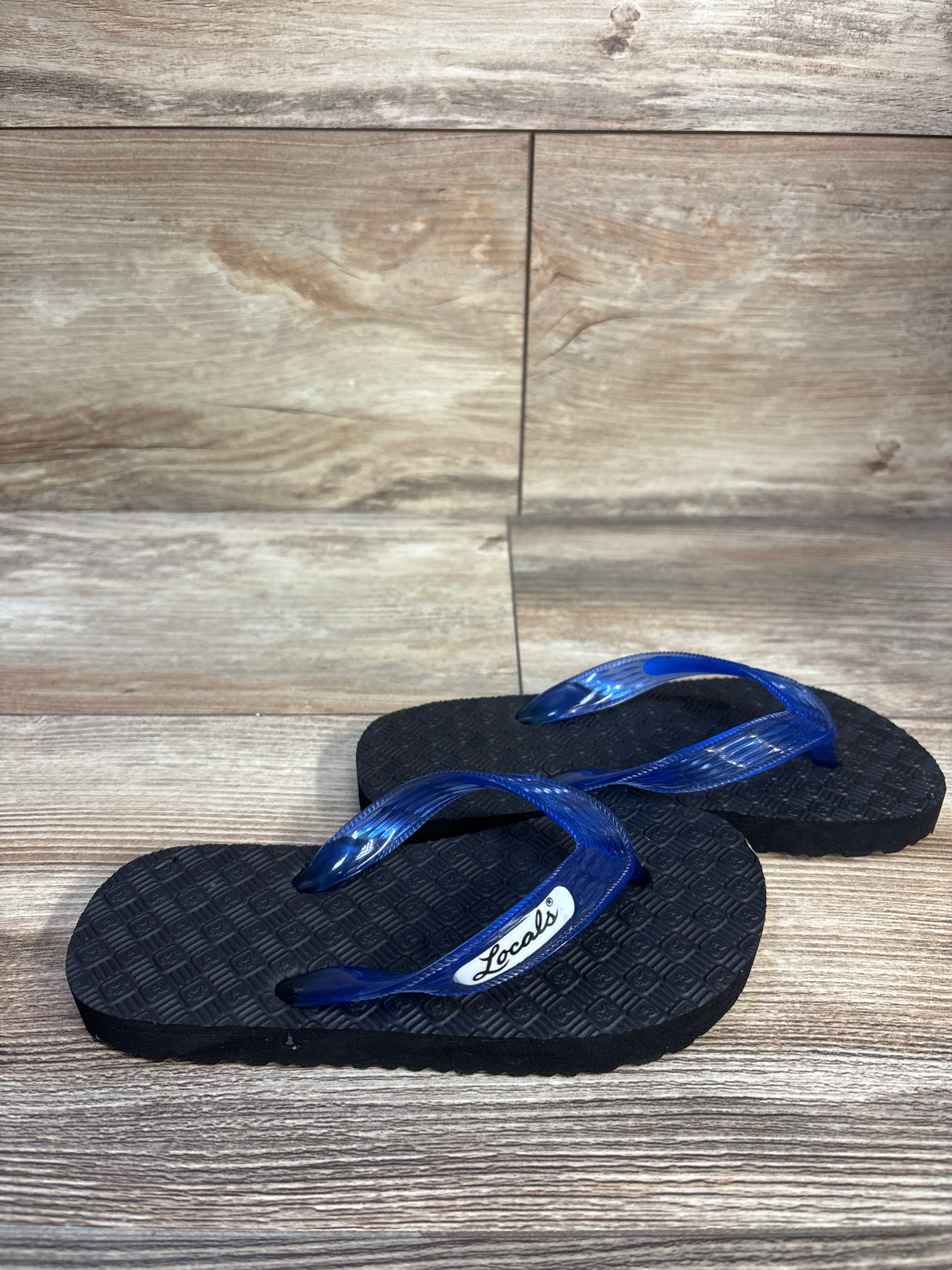 Angled view of Locals kids flip-flops size 7.5C showcasing translucent blue straps and textured black soles on wood