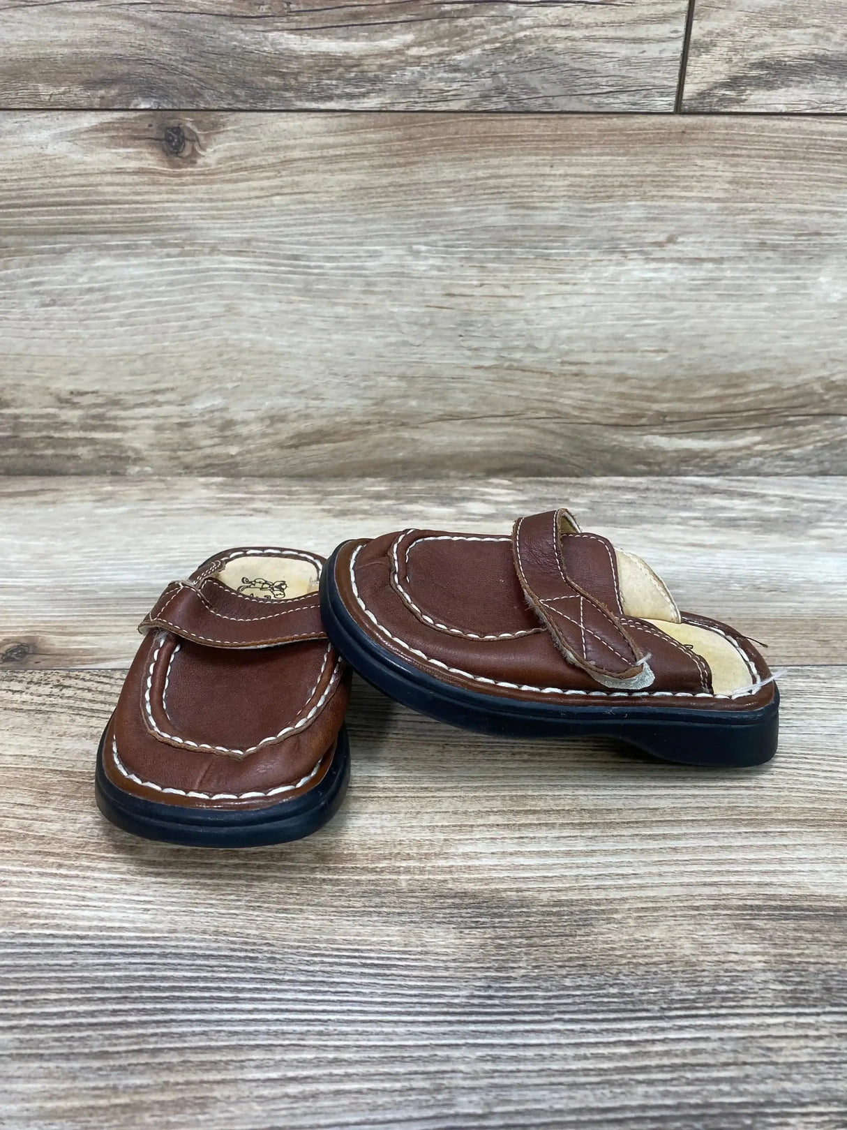 Three-quarter view of Tom Dog leather clogs in brown showing stitched upper, Velcro strap, and black sole on wood background