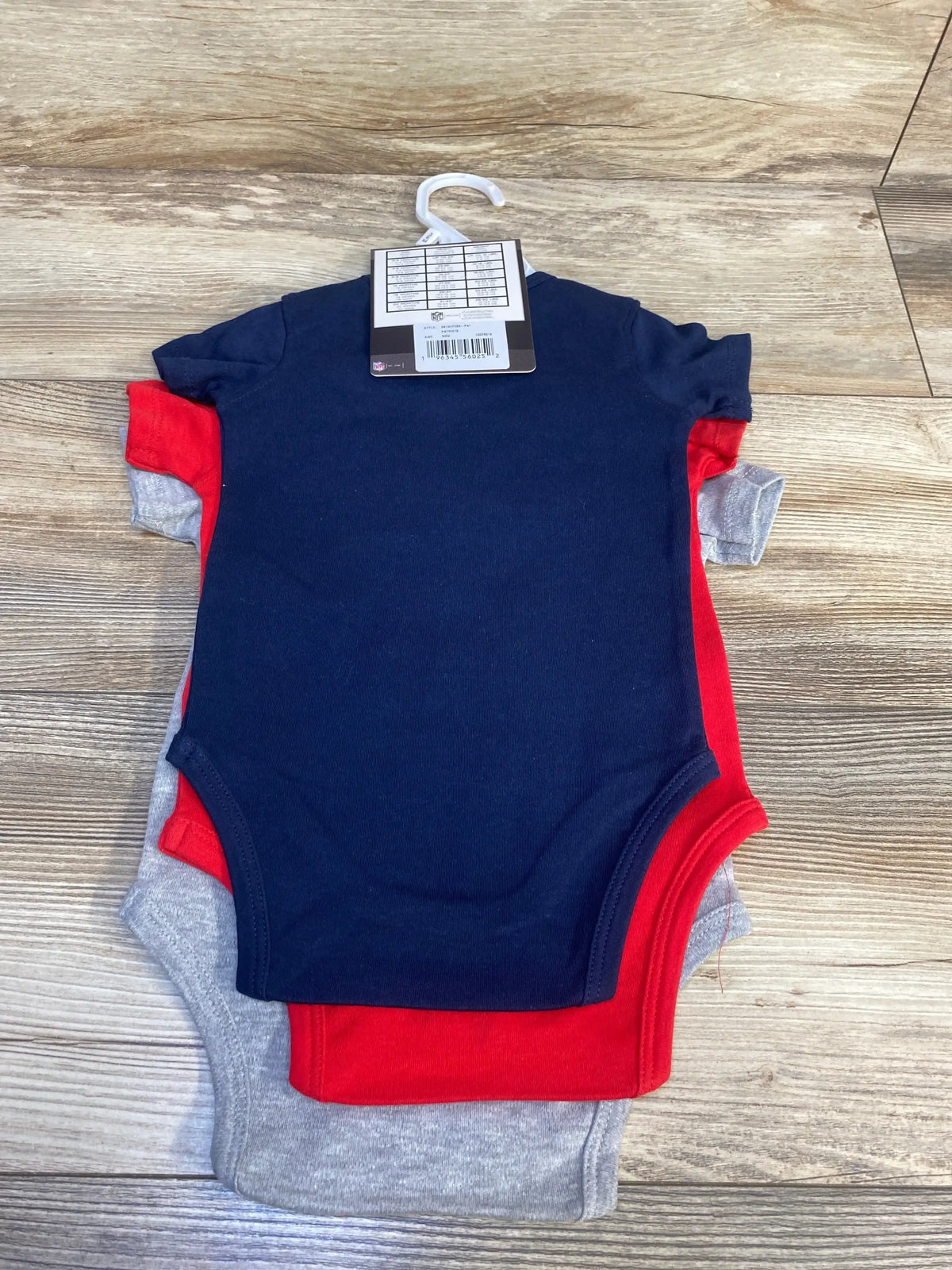Top view of 3-pack Patriots infant bodysuits in navy, red and gray showing back of navy piece with hanger and tag on wood background
