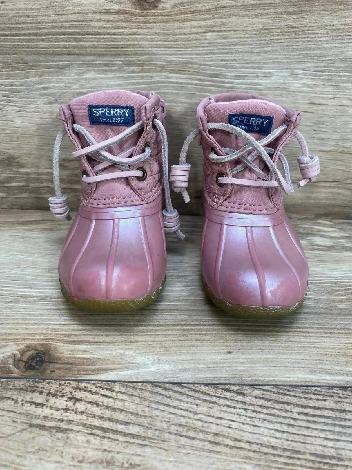 Front view of pink Sperry Saltwater toddler duck boots showing rubber toe lace up cords on wooden background