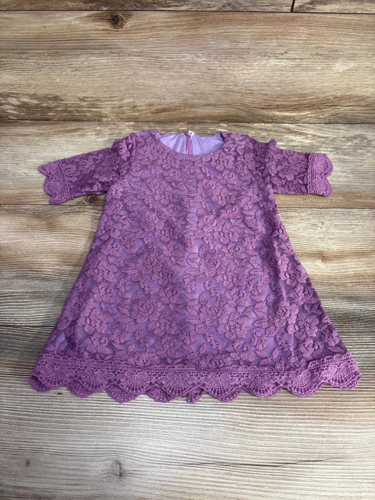 Flat lay of purple baby lace dress (12-18m) showing 3/4 sleeves, floral lace overlay, scalloped hem, and back zipper on wood background